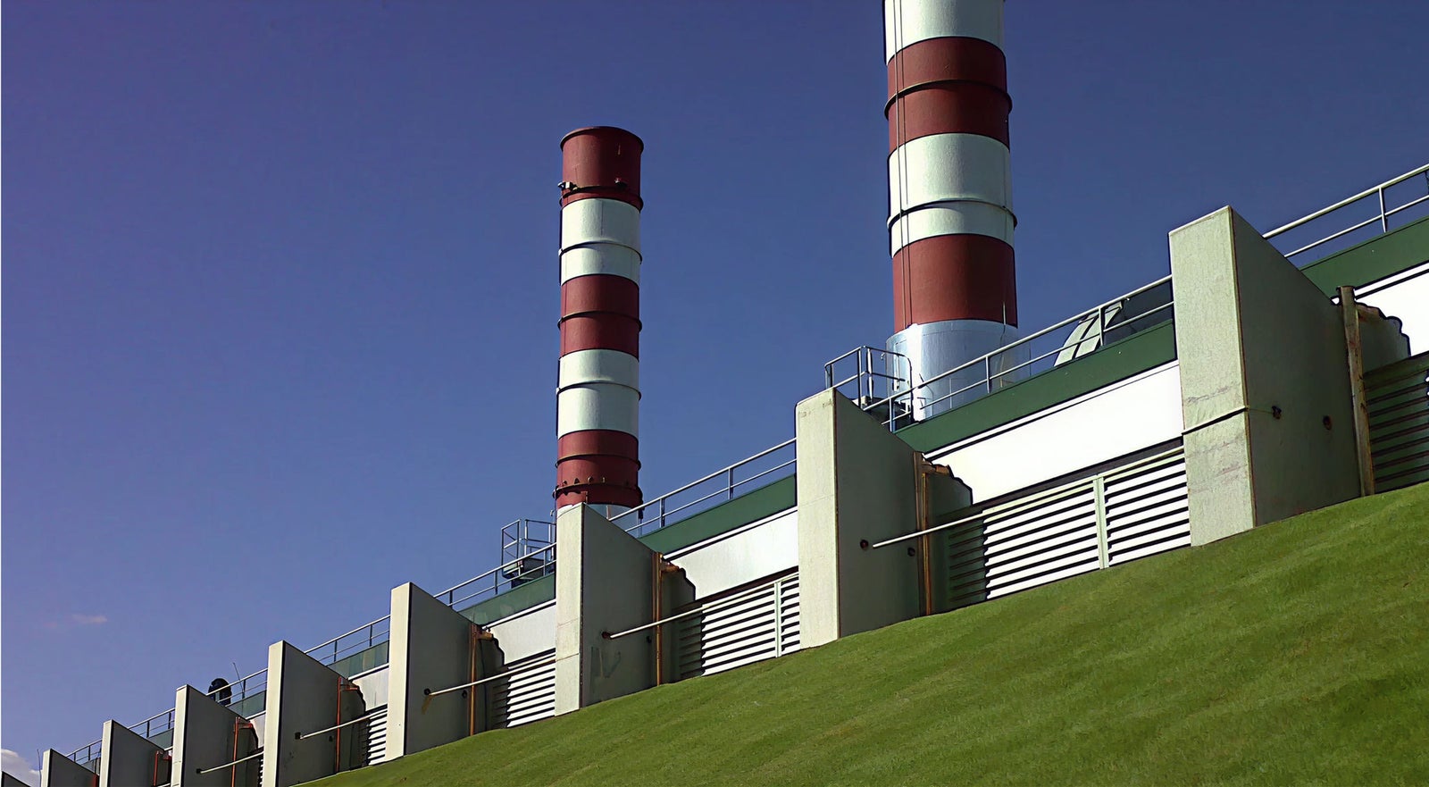 Fincantieri | Group - Subsidiaries and Affiliates - SOF: Large industrial facility featuring two tall chimneys with red and white stripes, clear blue sky and sloped green lawn in the foreground; modern industrial design with metallic panels and geometric shapes.