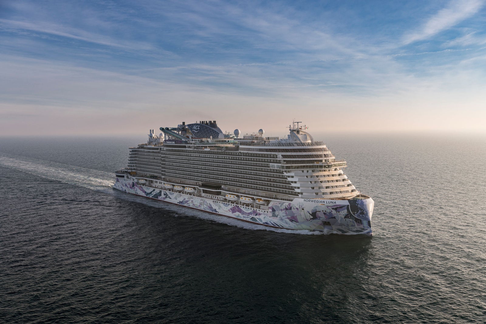 Fincantieri | Gruppo - Business - Prodotti - Navi da Crociera - Norwegian Luna: Nave da crociera moderna con design intricato blu e bianco in navigazione, su acque calme sotto cielo nuvoloso, più ponti con aree ricreative esterne, nome 'Norwegian Viva' visibile sulla prua.