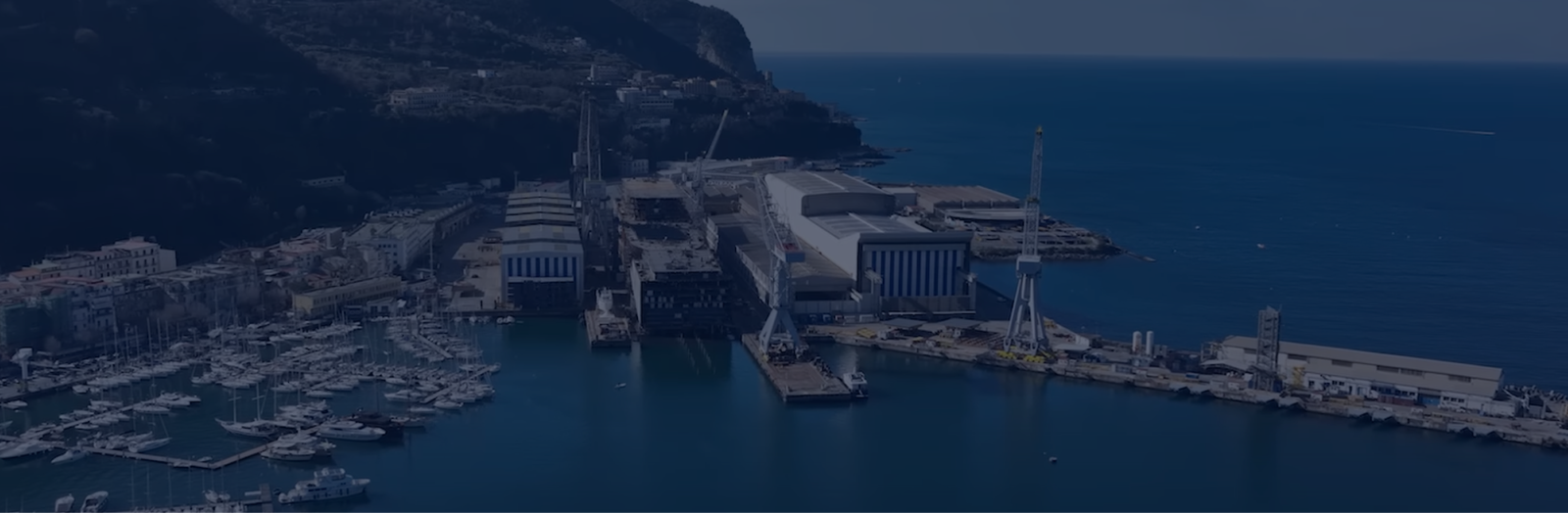 Fincantieri | Group - Innovation - Naval Ecosystem of the Future - Propulsion Systems and Clean Fuels: Large shipyard on a scenic coast with industrial buildings, cranes, and marina with docked boats; background of hills and open sea, blending industrial and nautical elements in a serene context.