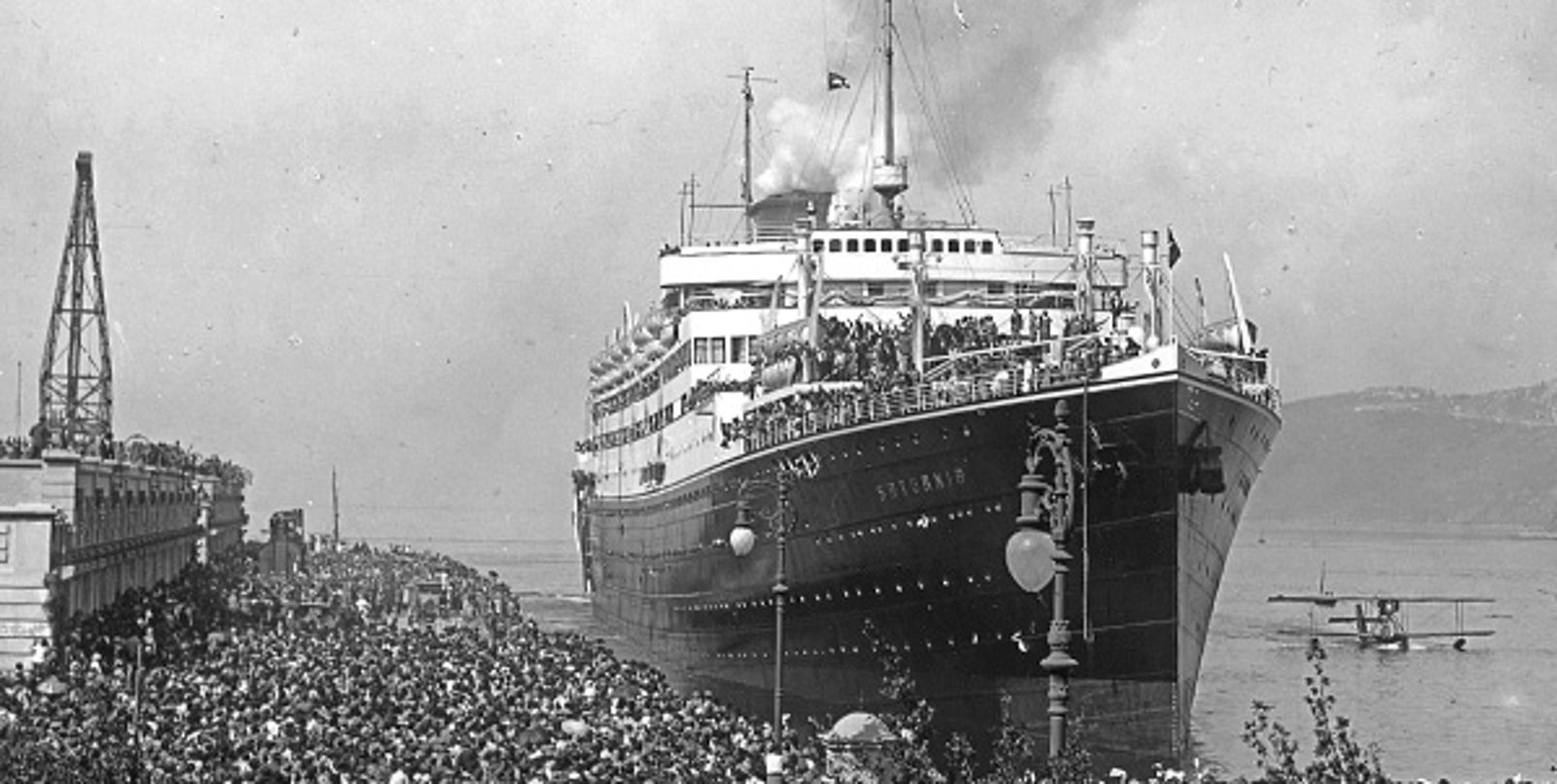 Fincantieri | About Us - Historic Ships: Large passenger ship 'Saturnia' arriving in a crowded port, large crowd on the quay, industrial structures and a seaplane in the background, smoke from the funnel highlighting activity, historic maritime scene.
