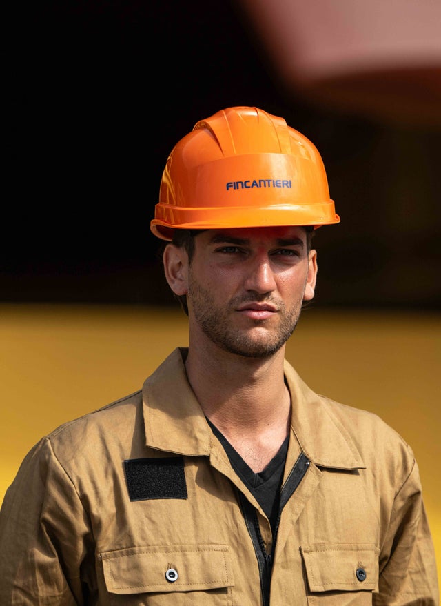 Fincantieri | Group - Sustainability - Industrial Excellence - Health and Safety: Person with orange Fincantieri helmet and brown work uniform, in an industrial or shipyard setting with blurred background, Fincantieri logo clearly visible on the helmet.