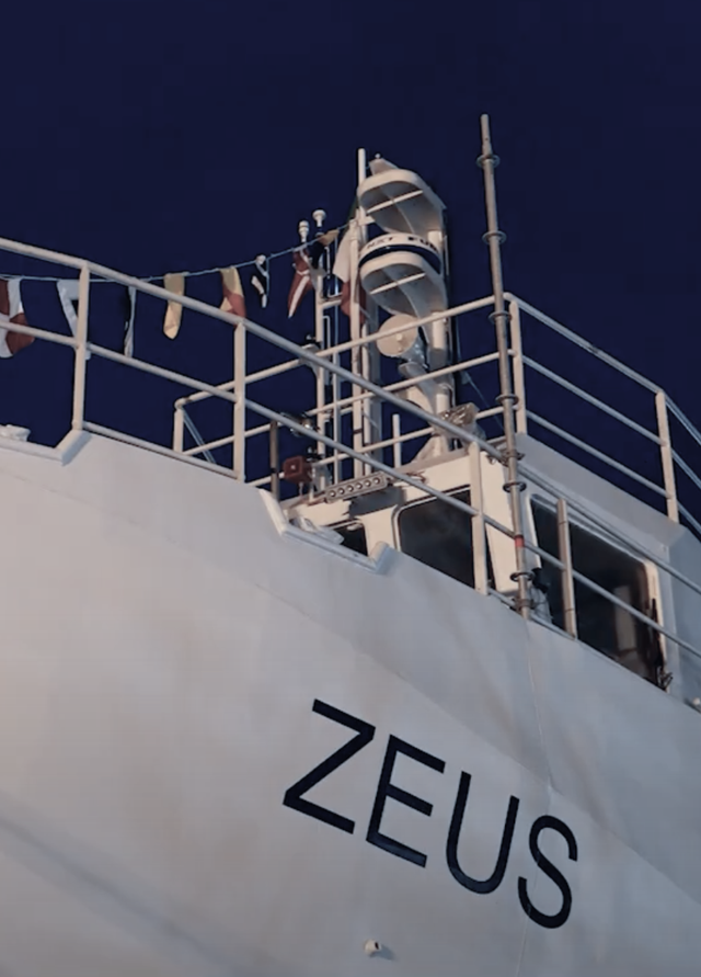Fincantieri | Group - Innovation - Naval Ecosystem of the Future - Propulsion Systems and Clean Fuels: White ship with the name ‘ZEUS’ clearly visible on the hull, railings, mast, and nautical flags, lighting highlighting the structure under a night sky.