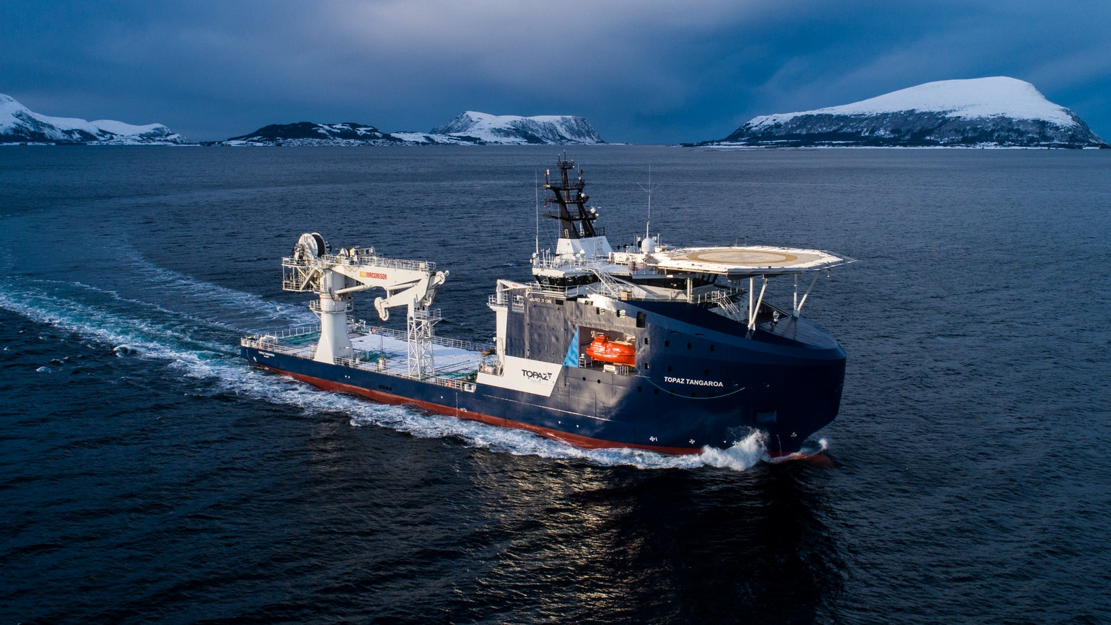 Prodotto - Offshore Support Vessel Topaz Tangaroa: Vista laterale della nave operativa in navigazione tra isole innevate, gru e parabole satellitari visibili sul ponte, ambiente artico orientato a esplorazione o operazioni marittime.