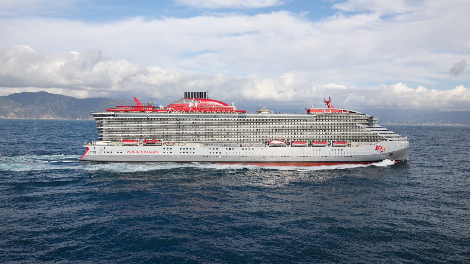 Fincantieri | Group - Business - Products - Cruise Ships - Valiant Lady: Modern cruise ship Virgin Voyages Scarlet Lady sailing on open waters under partly cloudy sky, sleek design with red accents and multiple decks, surrounding ocean and distant coastline create serene backdrop.