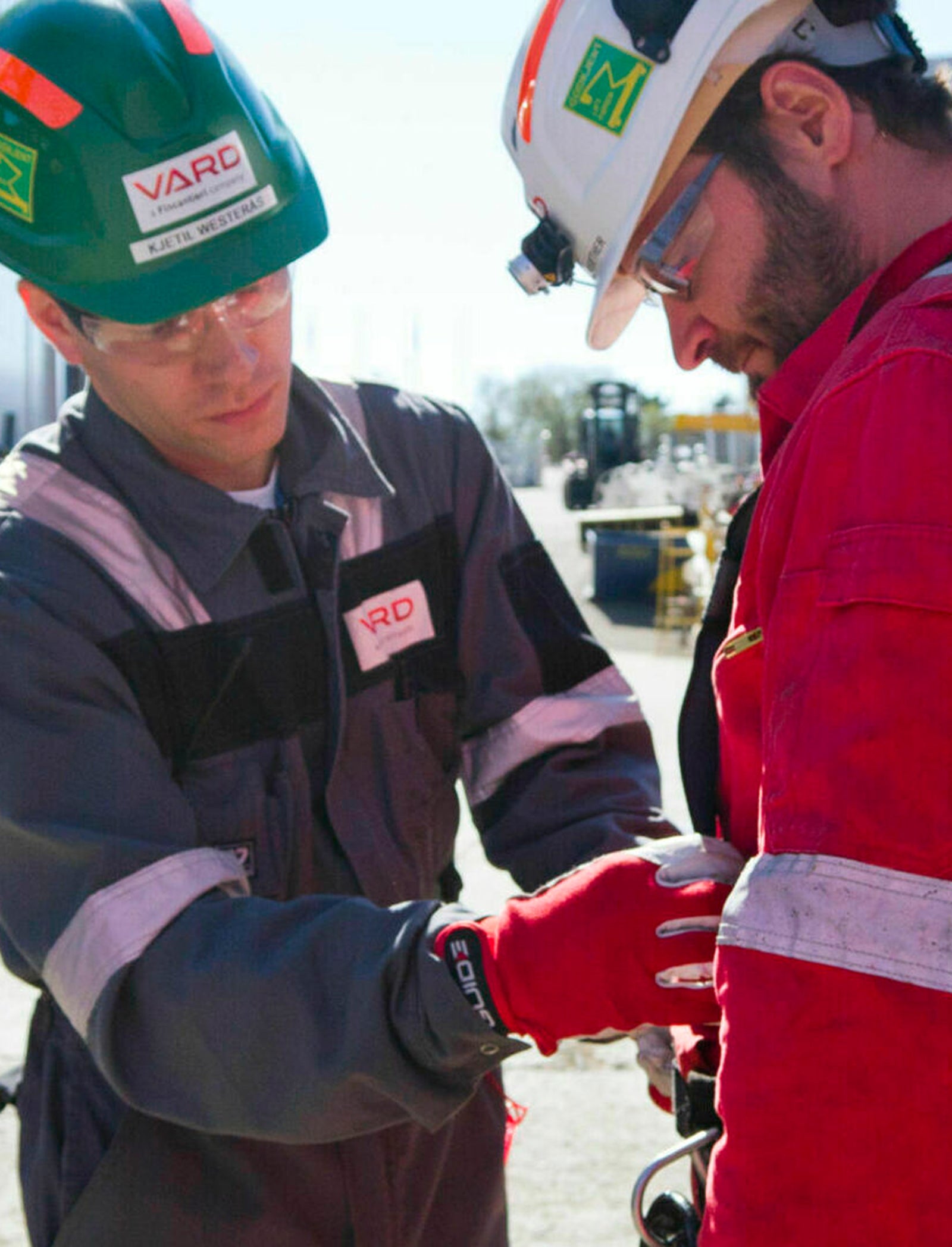 Fincantieri | Group - Subsidiaries and Affiliates - Vard Seaonics: Two industrial workers wearing protective helmets and uniforms in outdoor work setting, one in red uniform and the other in gray uniform with 'VARD' text visible; collaborative or safety-related activity, bright daylight and industrial equipment in the background.