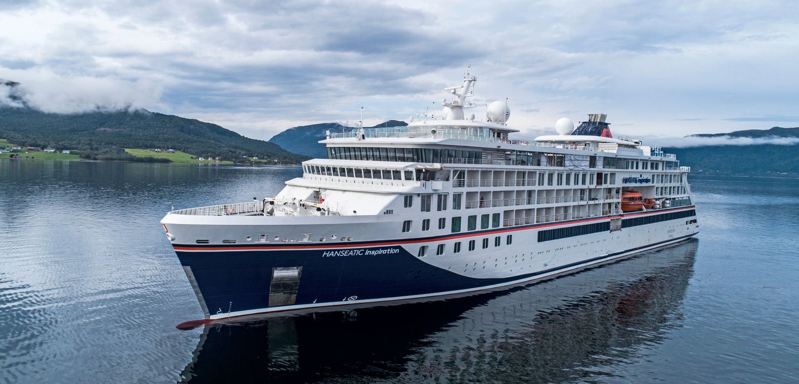 Fincantieri | Gruppo - Business - Prodotti - Navi da Crociera - Hanseatic Inspiration: Nave moderna operativa in navigazione in fiordo sereno, circondata da colline verdi e cielo nuvoloso, design elegante con accenti bianchi e blu navy, acque calme riflettono nave e paesaggio, atmosfera tranquilla.