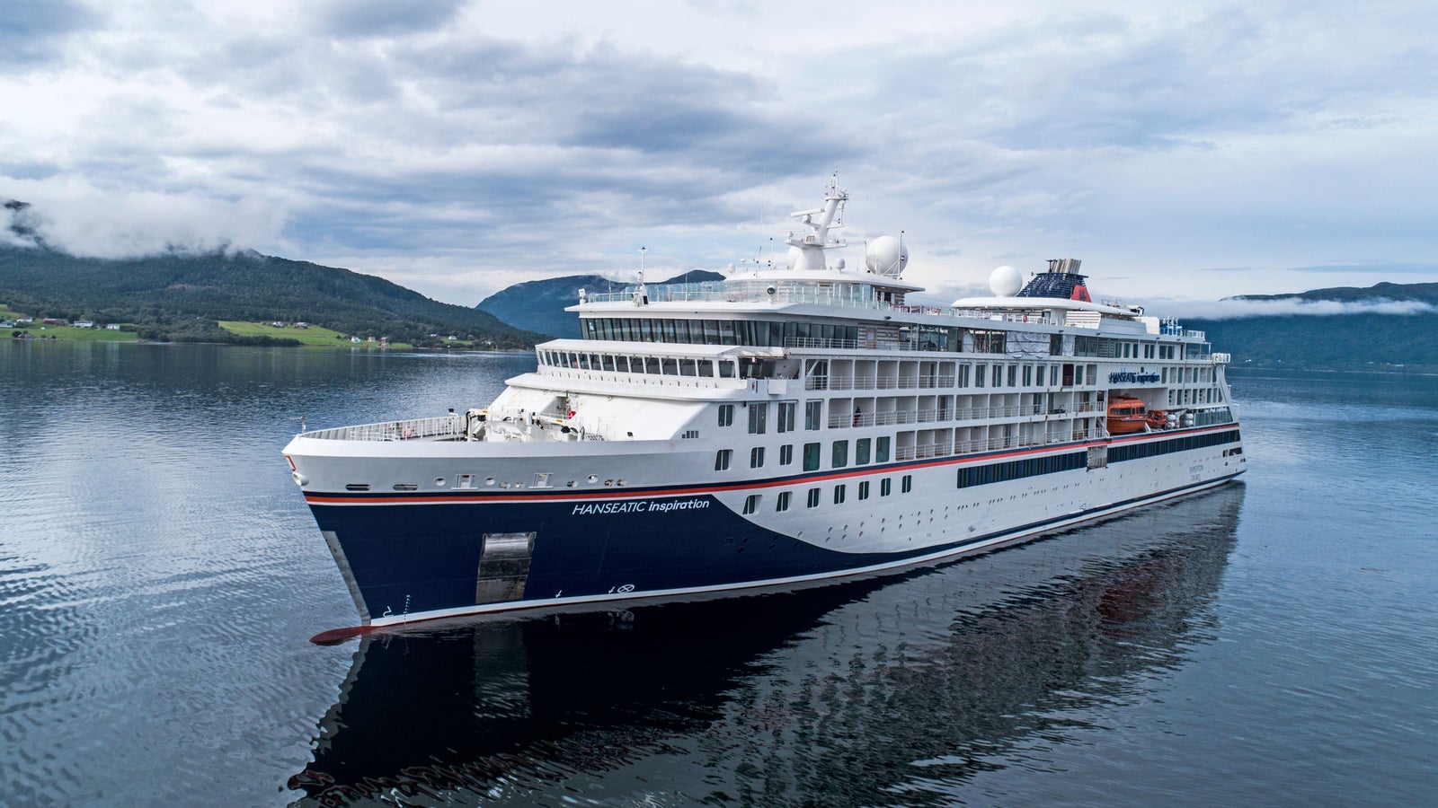 Fincantieri | Gruppo - Business - Prodotti - Navi da Crociera - Hanseatic Inspiration: Nave moderna operativa in navigazione in fiordo sereno, circondata da colline verdi e cielo nuvoloso, design elegante con accenti bianchi e blu navy, acque calme riflettono nave e paesaggio, atmosfera tranquilla.