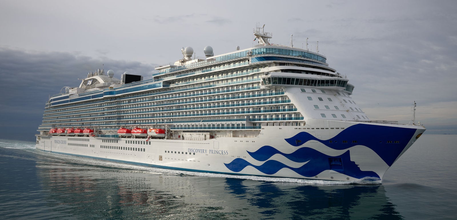 Fincantieri | Group - Business - Products - Cruise Ships - Discovery Princess: Large modern operational cruise ship sailing on calm waters under cloudy sky, multiple decks, red lifeboats and distinctive wave design on hull.