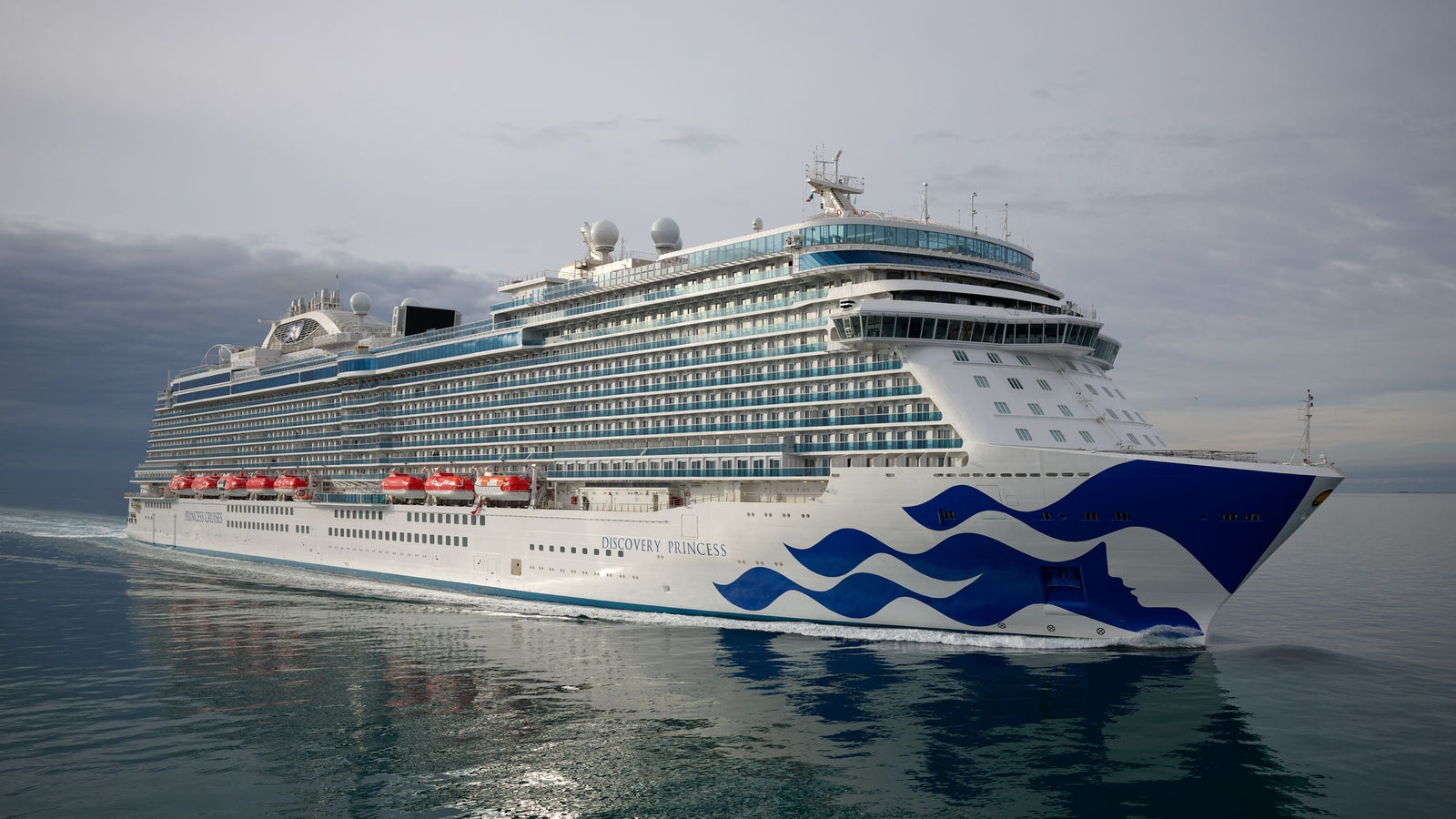Fincantieri | Group - Business - Products - Cruise Ships - Discovery Princess: Large modern operational cruise ship sailing on calm waters under cloudy sky, multiple decks, red lifeboats and distinctive wave design on hull.