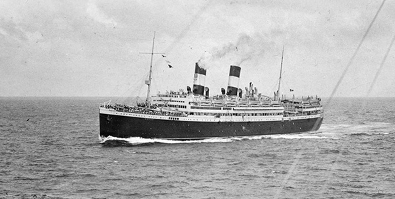 Fincantieri | About Us - Historic Ships: Large vintage ocean liner with two funnels sailing on open waters, classic maritime design, numerous passengers visible on the decks, cloudy sky and light waves against the hull.