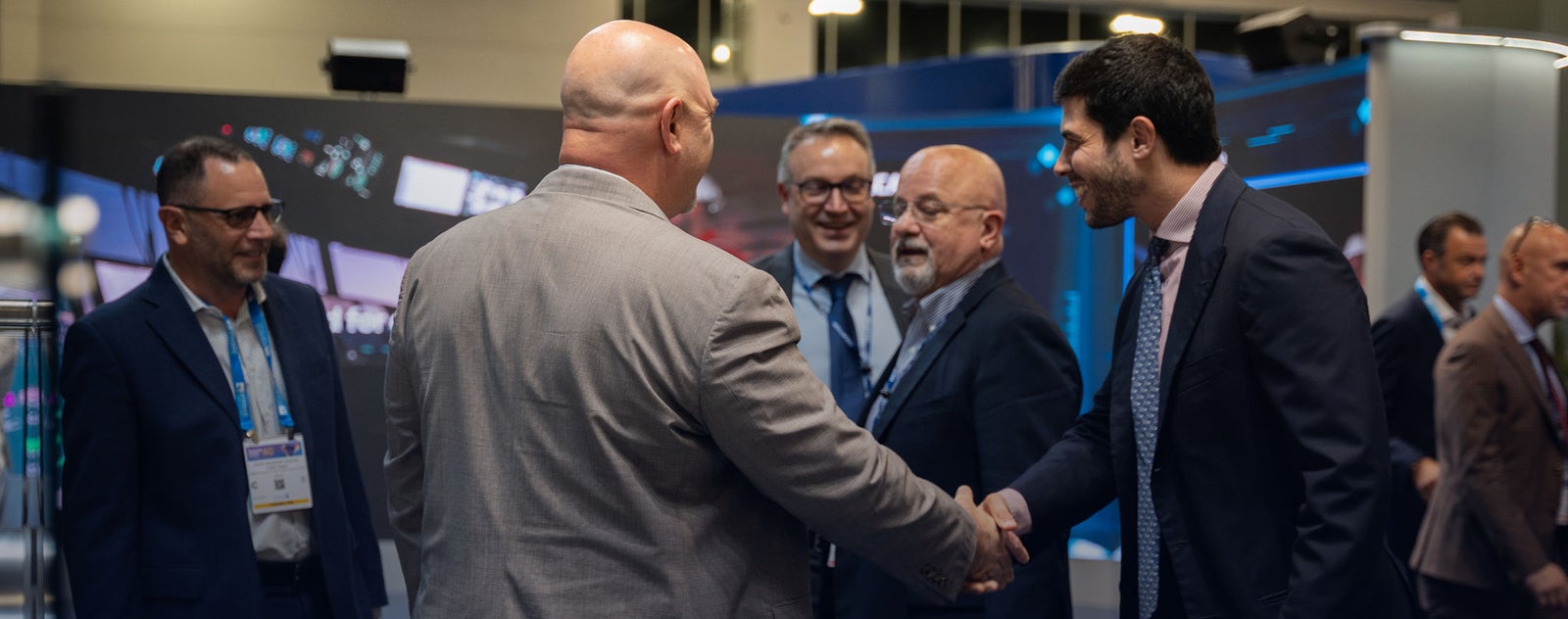Fincantieri | Gruppo - Sostenibilità - Eccellenza industriale - Cilenti: Gruppo di professionisti aziendali interagisce in conferenza corporate, ambiente moderno con schermi digitali e stand; due uomini si stringono la mano in gesto di networking o accordo, atmosfera formale con partecipanti in abiti eleganti.