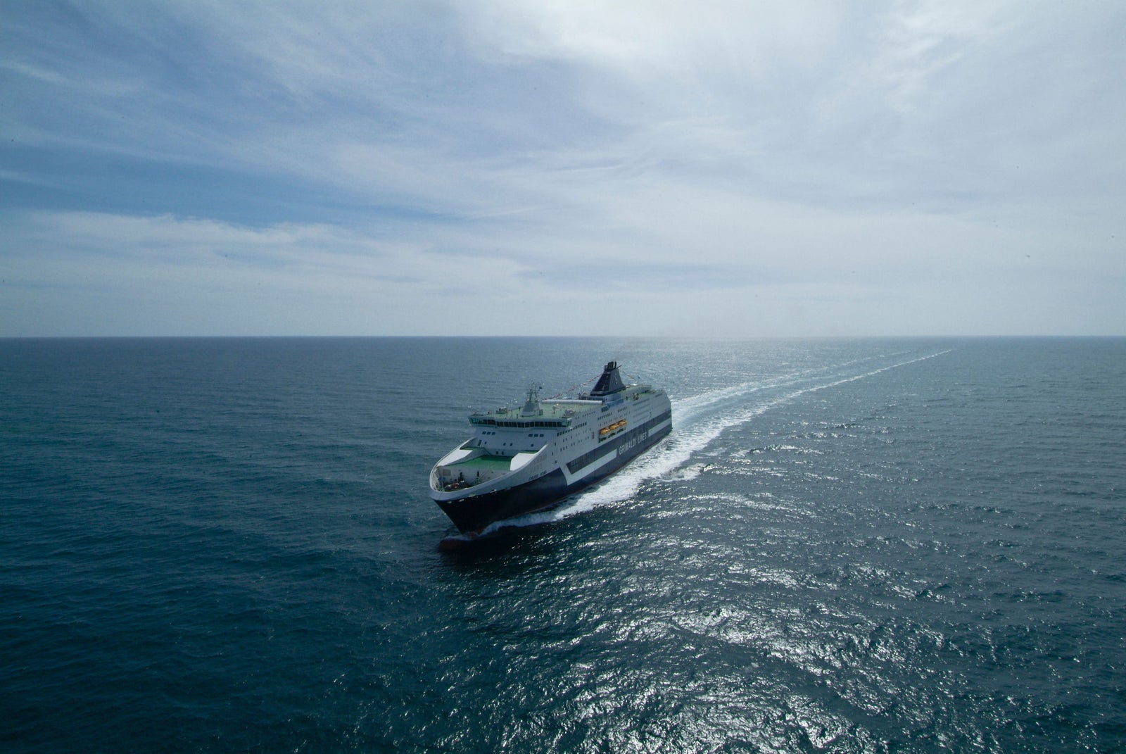 Prodotto - Traghetto Roma Grimaldi Lines: Vista laterale della nave operativa in navigazione, scia bianca evidente, design slanciato a più ponti, scafo verde e bianco con scritta GRIMALDI LINES ben visibile, ambiente marittimo soleggiato.