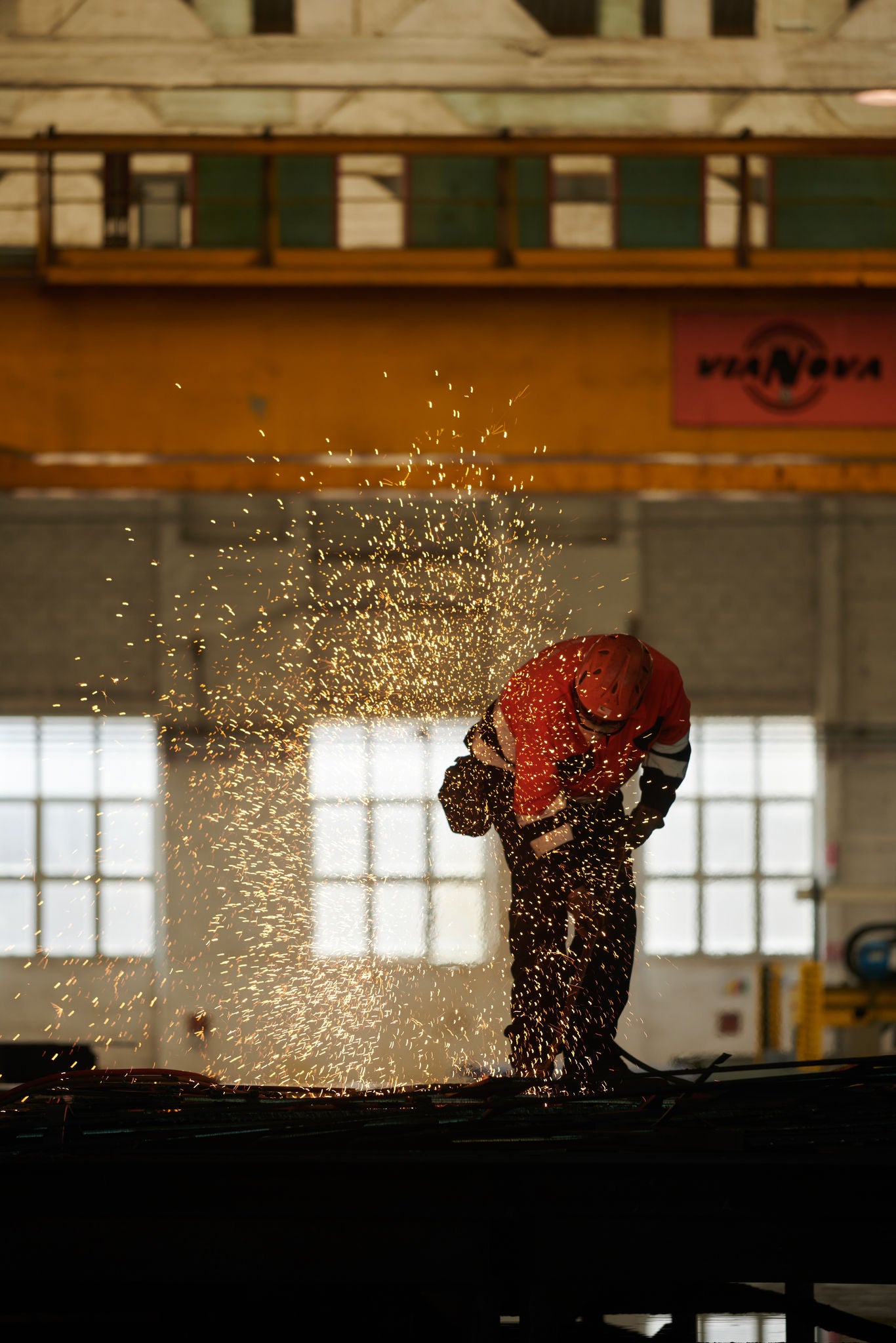 Fincantieri | Group - About Us - Goals and Strategy - Business Model: Worker with protective equipment welding metal in an industrial factory; bright sparks during the process, background with large windows and yellow crane bearing the text ‘Via Nova’.