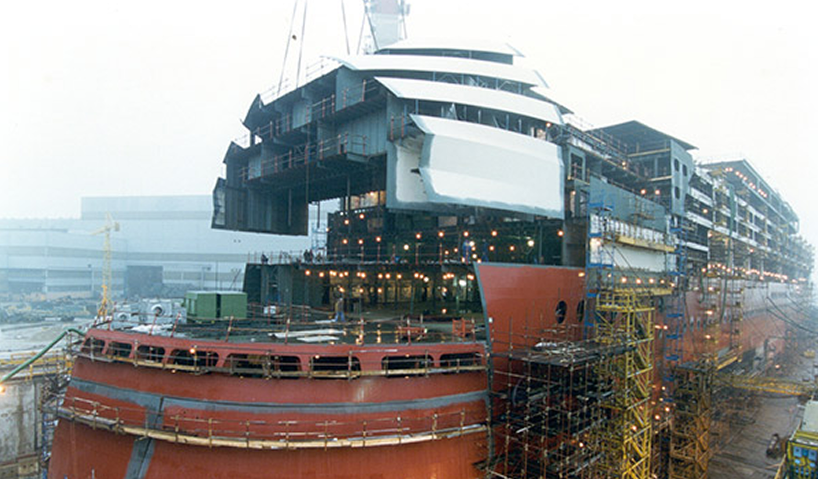 Fincantieri | About Us - Heritage and Future: Assembly of a large ship in an active shipyard, scaffolding and industrial equipment, partially completed hull and upper decks, workers and machinery visible, industrial atmosphere with overcast sky.