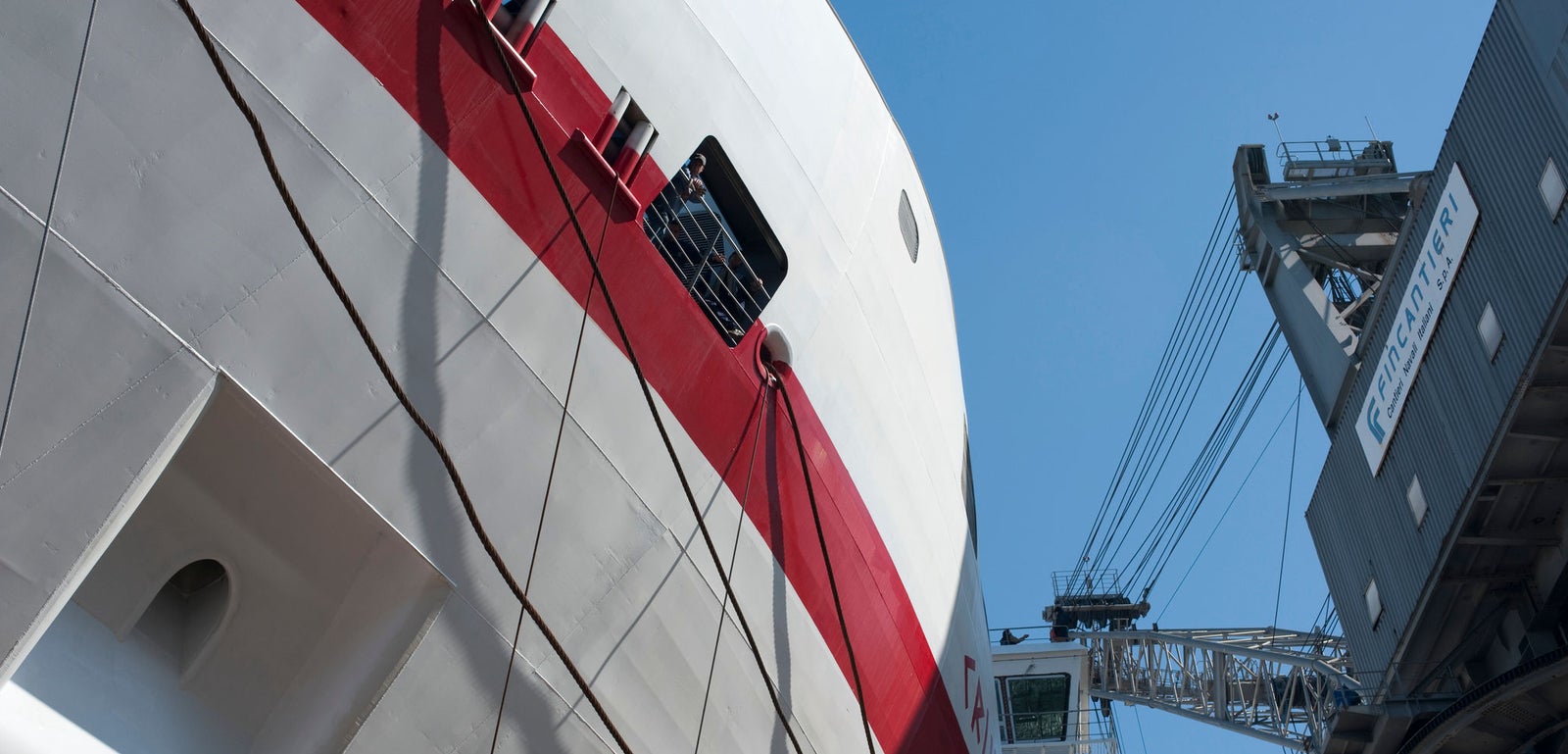 Fincantieri | Group - Business - Products - Ferries - Cruise Europa: Close-up view of large ship with red and white exterior docked near industrial crane; port or shipyard setting under clear blue skies; visible text 'MacGregor' on crane structure, enhancing maritime context.