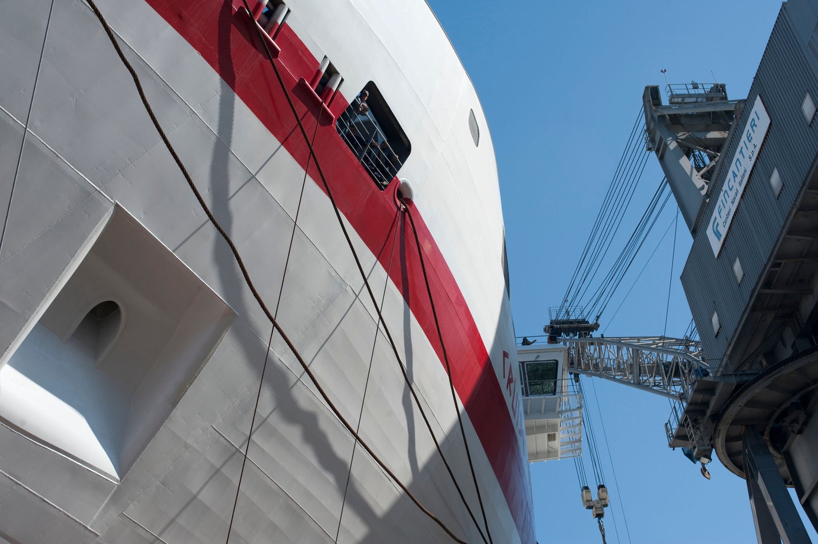 Prodotto - Traghetto Europa 8 Minoan Lines: Dettaglio nave ormeggiata con scafo bianco e rosso vicino a una gru industriale, scritta MacGregor visibile sulla gru, contesto portuale, cielo sereno.
