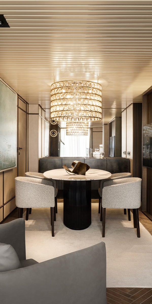 Fincantieri | Gruppo - Società - Controllate e collegate - Operae Interiors: Sala da pranzo contemporanea con tavolo tondo nero e sedie imbottite in palette neutra, illuminazione da chandelier in cristallo che aggiunge eleganza; pannelli in legno e tappeto soffice, atmosfera calda e accogliente.