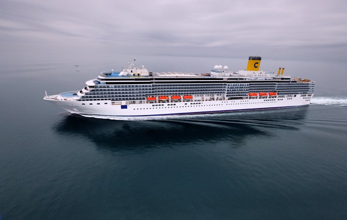 Fincantieri | Group - Business - Products - Cruise Ships - Costa Deliziosa: Massive cruise ship in operation gliding through calm open waters under cloudy sky, multiple decks, lifeboats, distinctive yellow funnel with visible 'C' logo, serene ocean setting emphasizes ship's grandeur and scale.