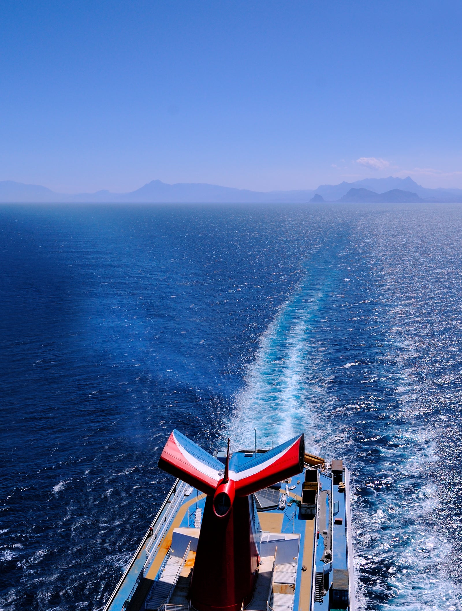 Fincantieri | Group - Business - Products - Cruise Ships - Carnival Splendor operativa: Large operational cruise ship sailing in the open ocean, visible wake behind, clear blue sky, distant islands on the horizon, prominent blue wave design on hull stands out against serene marine landscape.