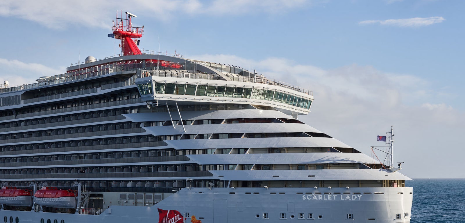 Fincantieri | Group - Business - Products - Cruise Ships - Scarlet Lady: Side view of Scarlet Lady cruise ship bow in operation, modern ship with multiple decks, distinctive red funnel, 'Virgin' logo and 'Scarlet Lady' name clearly visible on hull, sailing on calm waters under partly cloudy sky.