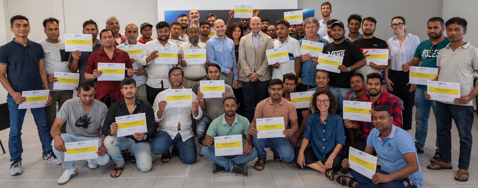 Sustainability - People and Communities - Training Event: Large group of men and women of various ages holding yellow and white certificates, indoor environment, blue banner in the background. People arranged in rows, sitting and standing.
