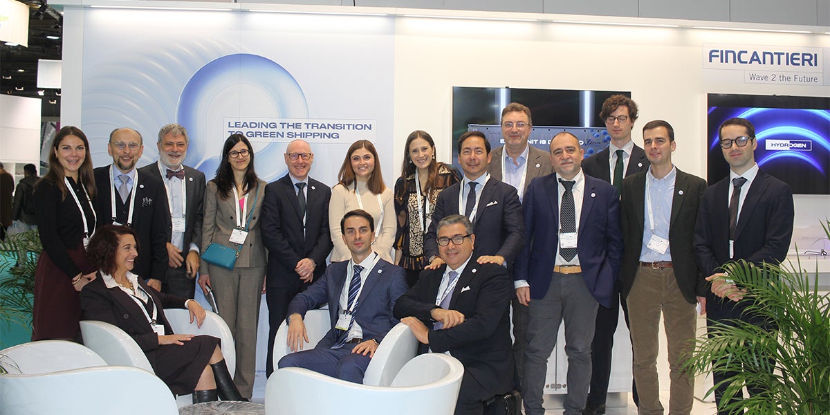 Fincantieri | Group - Innovation - Naval Ecosystem of the Future - Wave 2 the Future: Group of people posing at a professional event at the Fincantieri stand, visible texts on green shipping and sustainability, modern furnishings and plants for a corporate and eco-friendly theme.