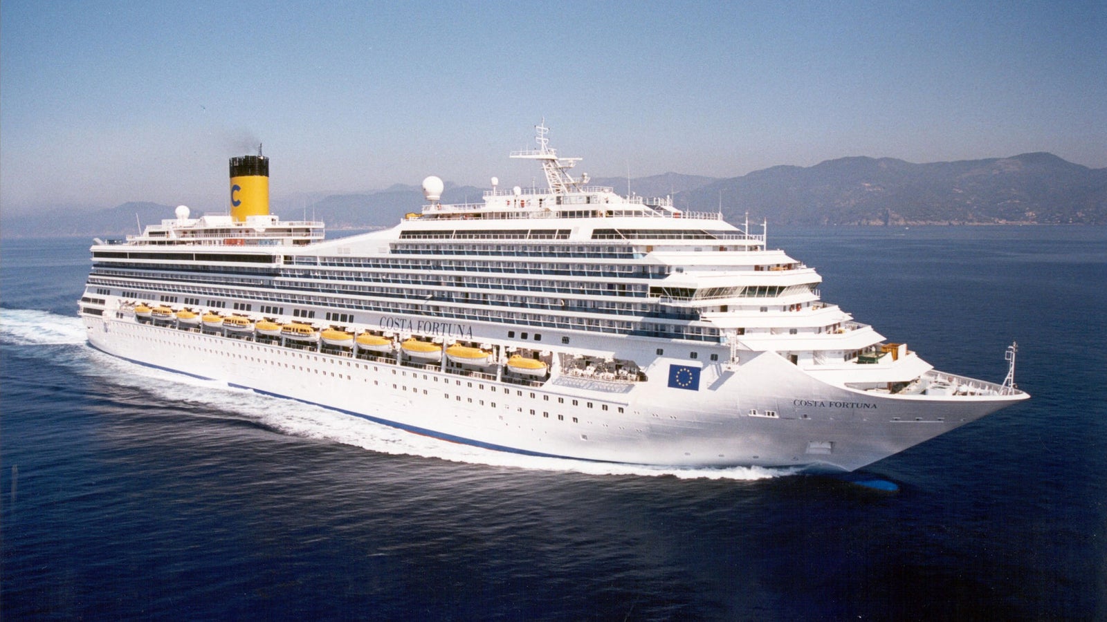 Fincantieri | Group - Business - Products - Cruise Ships - Costa Fortuna: Side view of Costa Fortuna cruise ship in operation, white hull with multiple passenger decks, visible yellow lifeboats, funnel with Costa logo, mountainous profile in the background.