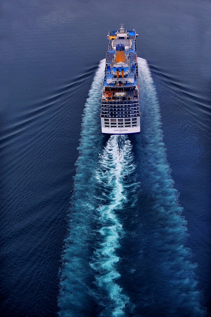  Fincantieri | Group - Business - Products - Cruise Ships - Costa Deliziosa: Aerial view of massive cruise ship in operation sailing through calm deep blue waters, striking white wake behind emphasizes movement and scale, serene ocean voyage setting with no land visible.