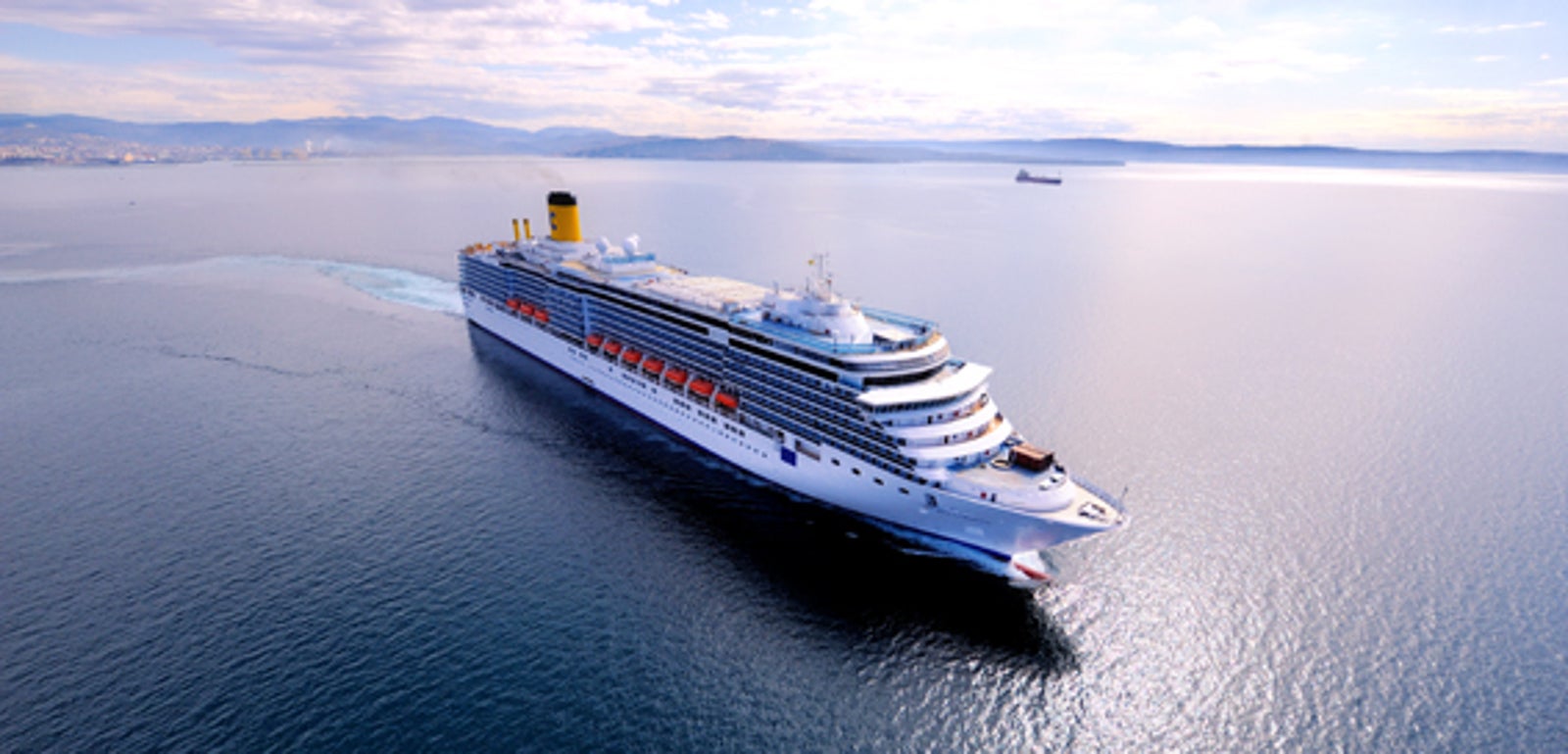  Fincantieri | Gruppo - Business - Prodotti - Navi da Crociera - Costa Luminosa: Grande nave da crociera operativa con molti ponti e scialuppe di salvataggio visibili lungo i lati, scivola su acque calme sotto cielo luminoso, design elegante e moderno.