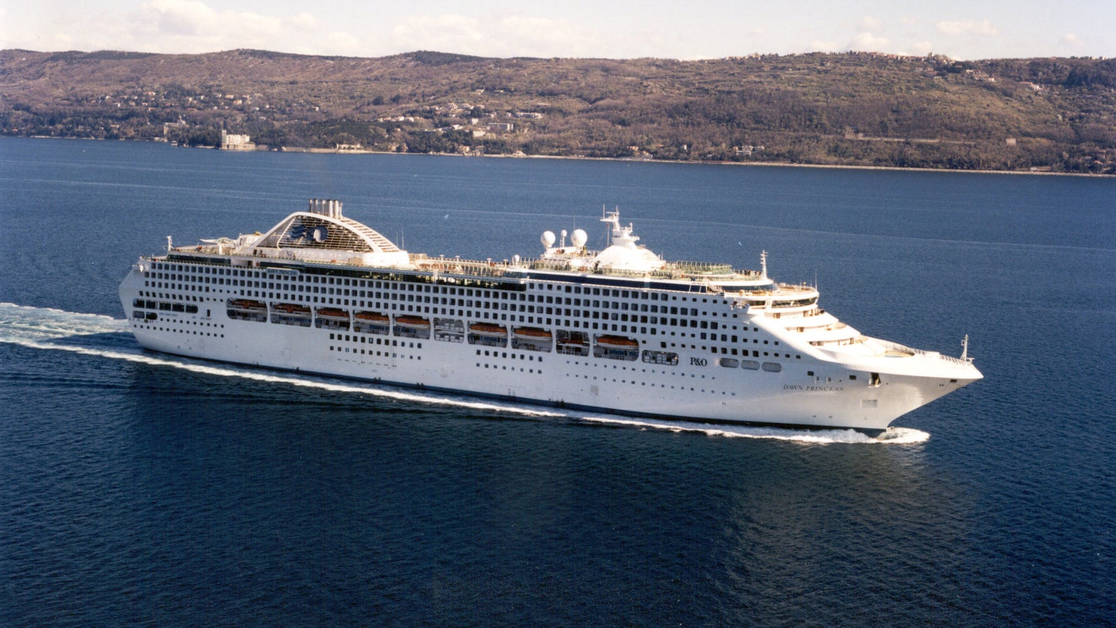 Fincantieri | Group - Business - Products - Cruise Ships - Dawn Princess: Side view of Dawn Princess cruise ship in operation navigating, white hull, multiple decks, upper dome, coastal background.