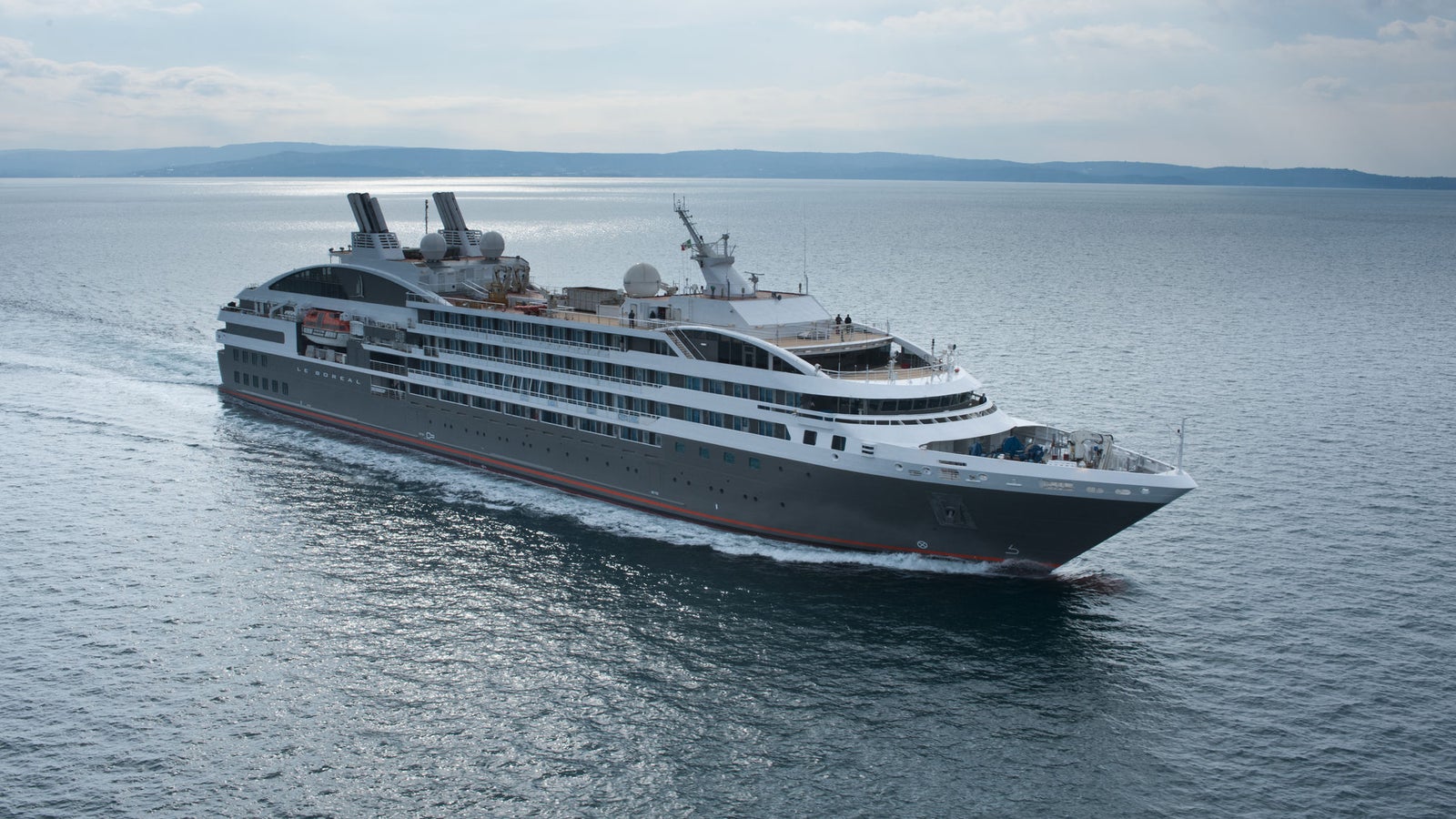Fincantieri | Group - Business - Products - Cruise Ships - Le Boreal: Side view of Le Boreal cruise ship in operation navigating open sea, dark linear profile hull, multiple passenger decks, partly cloudy sky in background.