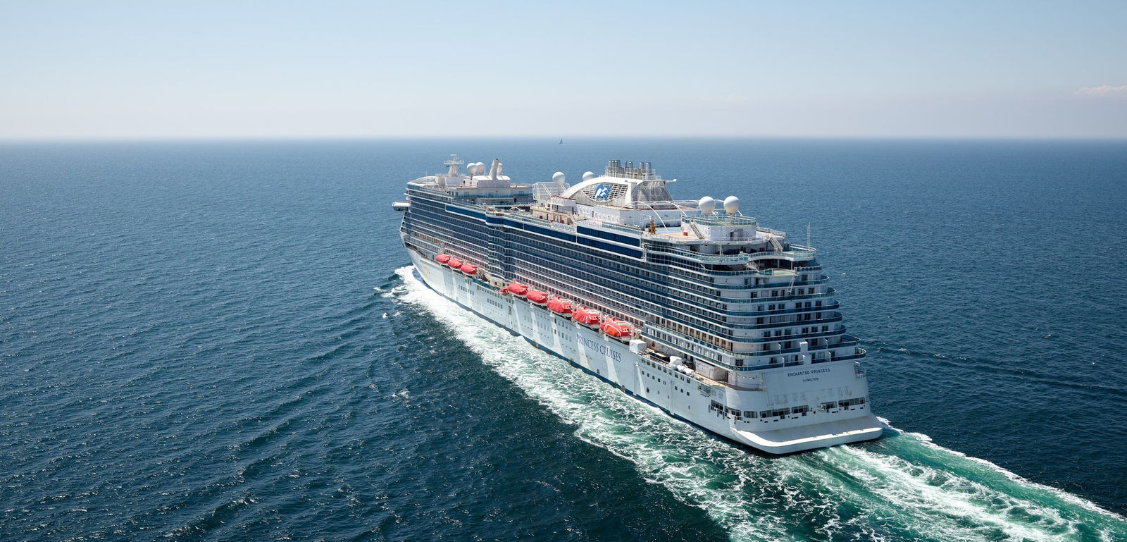 Fincantieri | Group - Business - Products - Cruise Ships - Enchanted Princess: Overhead stern view of Enchanted Princess cruise ship in operation, gliding on calm ocean waters under clear blue sky, elegant design with wave patterns on hull.