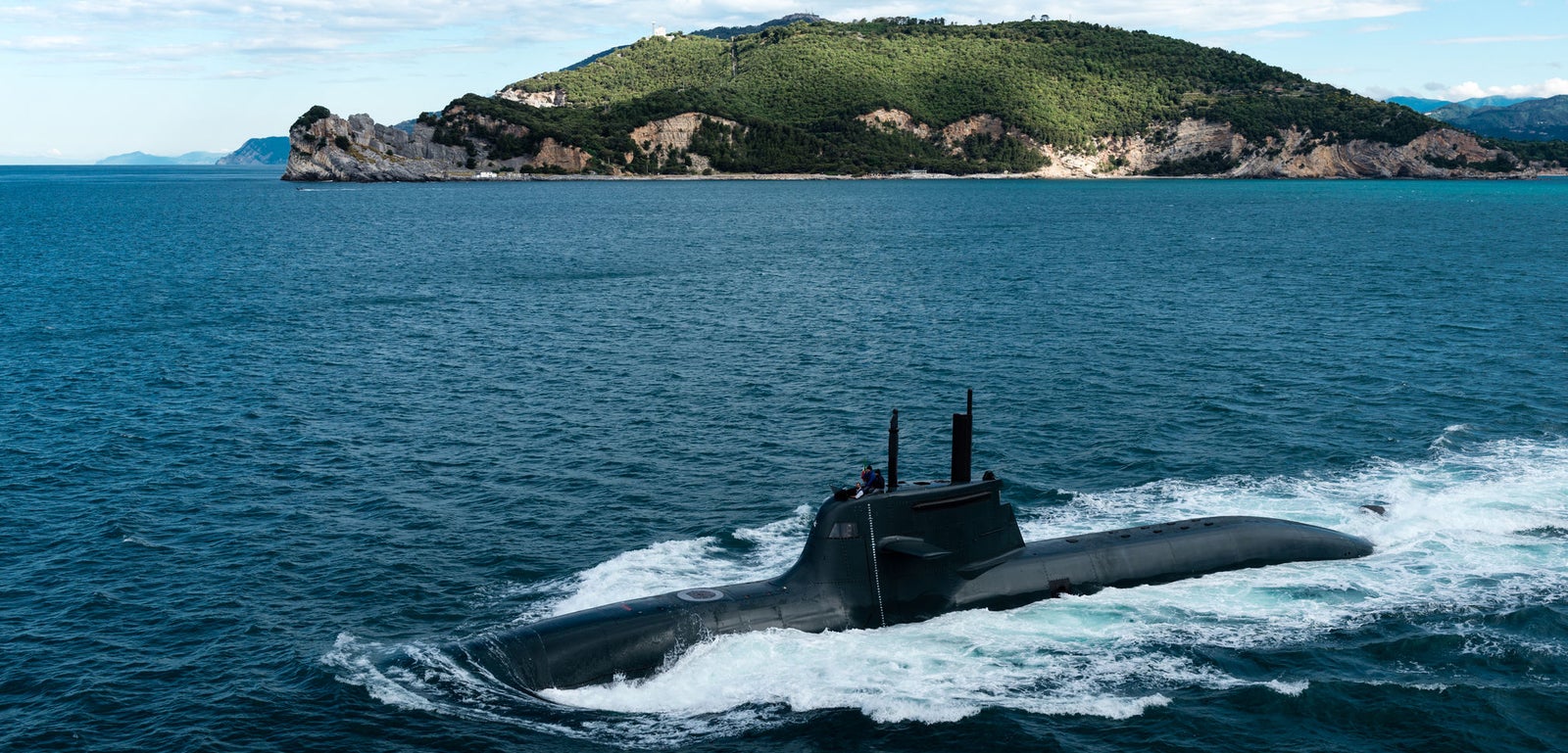 Fincantieri | Gruppo - Business - Prodotti - Sommergibili - Classe Todaro: Sommergibile nero slanciato operativo, in navigazione su acque blu calme vicino a un’isola costiera verdeggiante, terreno roccioso e vegetazione visibili, scia dolce, cielo parzialmente nuvoloso.