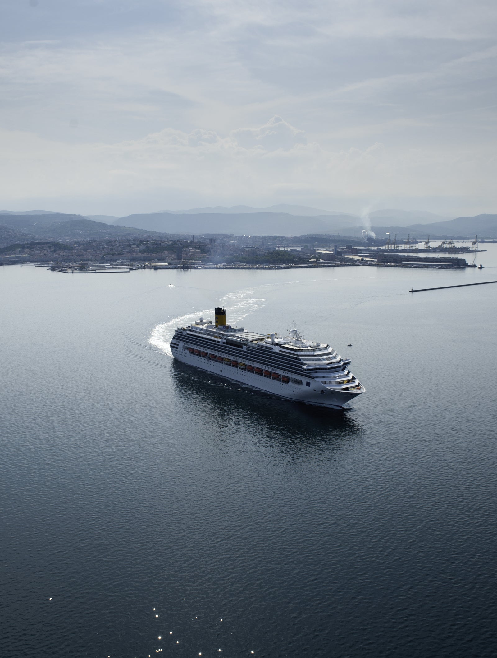 Fincantieri | Group - Business - Products - Cruise Ships - Costa Favolosa: Large white cruise ship in operation sailing in calm open waters near coastal port, multiple decks and visible lifeboats along sides, background with industrial structures, distant hills and hazy sky, scene conveys travel and leisure.