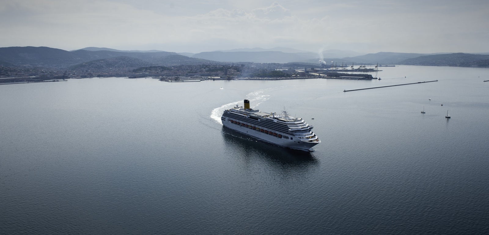Fincantieri | Group - Business - Products - Cruise Ships - Costa Favolosa: Large white cruise ship in operation sailing in calm open waters near coastal port, multiple decks and visible lifeboats along sides, background with industrial structures, distant hills and hazy sky, scene conveys travel and leisure.