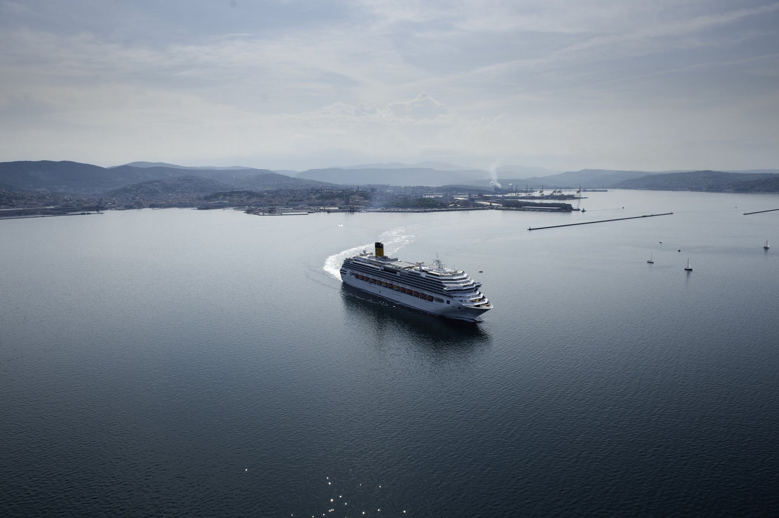 Fincantieri | Group - Business - Products - Cruise Ships - Costa Favolosa: Large white cruise ship in operation sailing in calm open waters near coastal port, multiple decks and visible lifeboats along sides, background with industrial structures, distant hills and hazy sky, scene conveys travel and leisure.