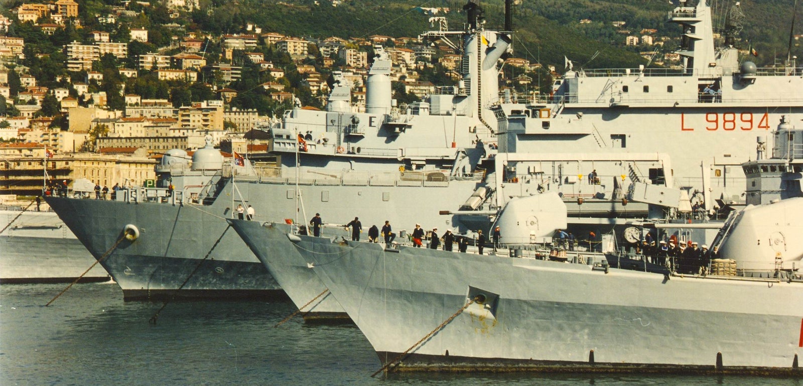 Fincantieri | Gruppo - Business - Prodotti - Navi Militari - Portaerei e Navi Anfibi - Classe San Giusto: Gruppo di navi militari ormeggiate in porto costiero, numeri identificativi visibili tra cui "L9894", persone che camminano sui ponti, vista città, colline e edifici residenziali sullo sfondo.