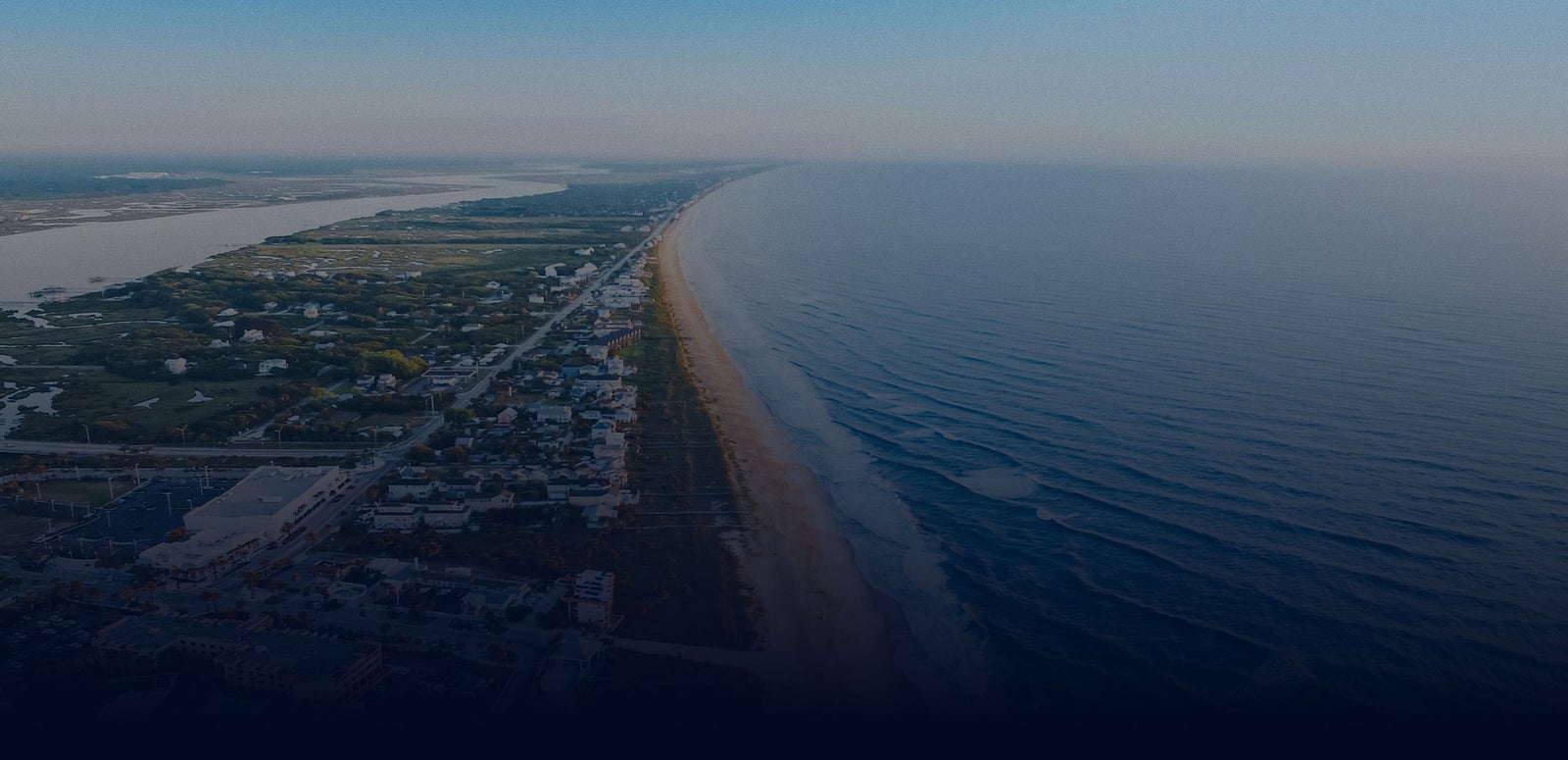 Fincantieri | Group - Sustainability - Energy Transition - Respect for Biodiversity: Aerial view of coastal area with a long curved beach along the shoreline, urban or residential area visible inland and ocean stretching to the horizon under an overcast sky.