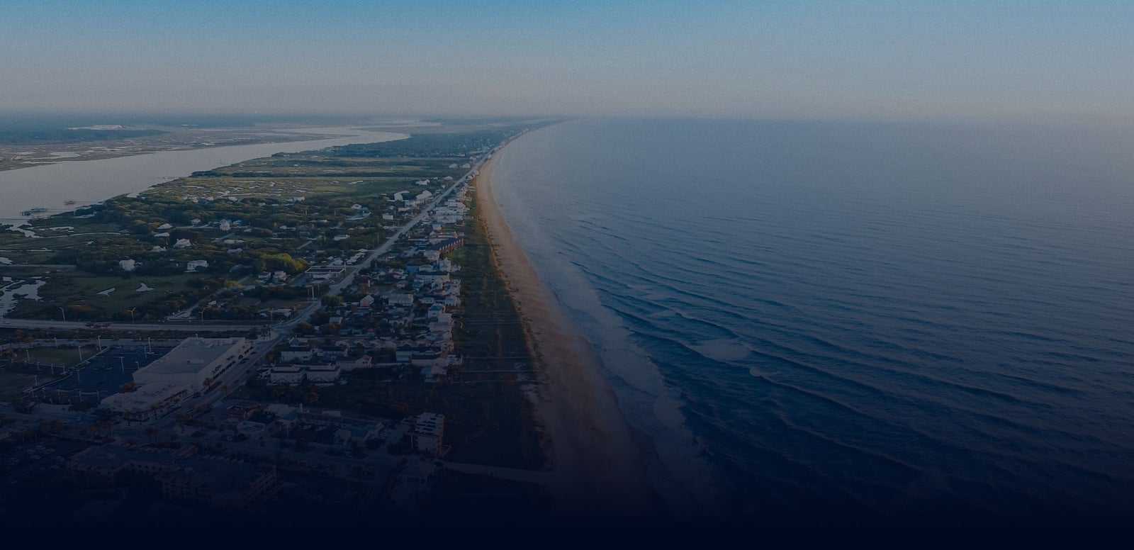 Fincantieri | Group - Sustainability - Energy Transition - Respect for Biodiversity: Aerial view of coastal area with a long curved beach along the shoreline, urban or residential area visible inland and ocean stretching to the horizon under an overcast sky.