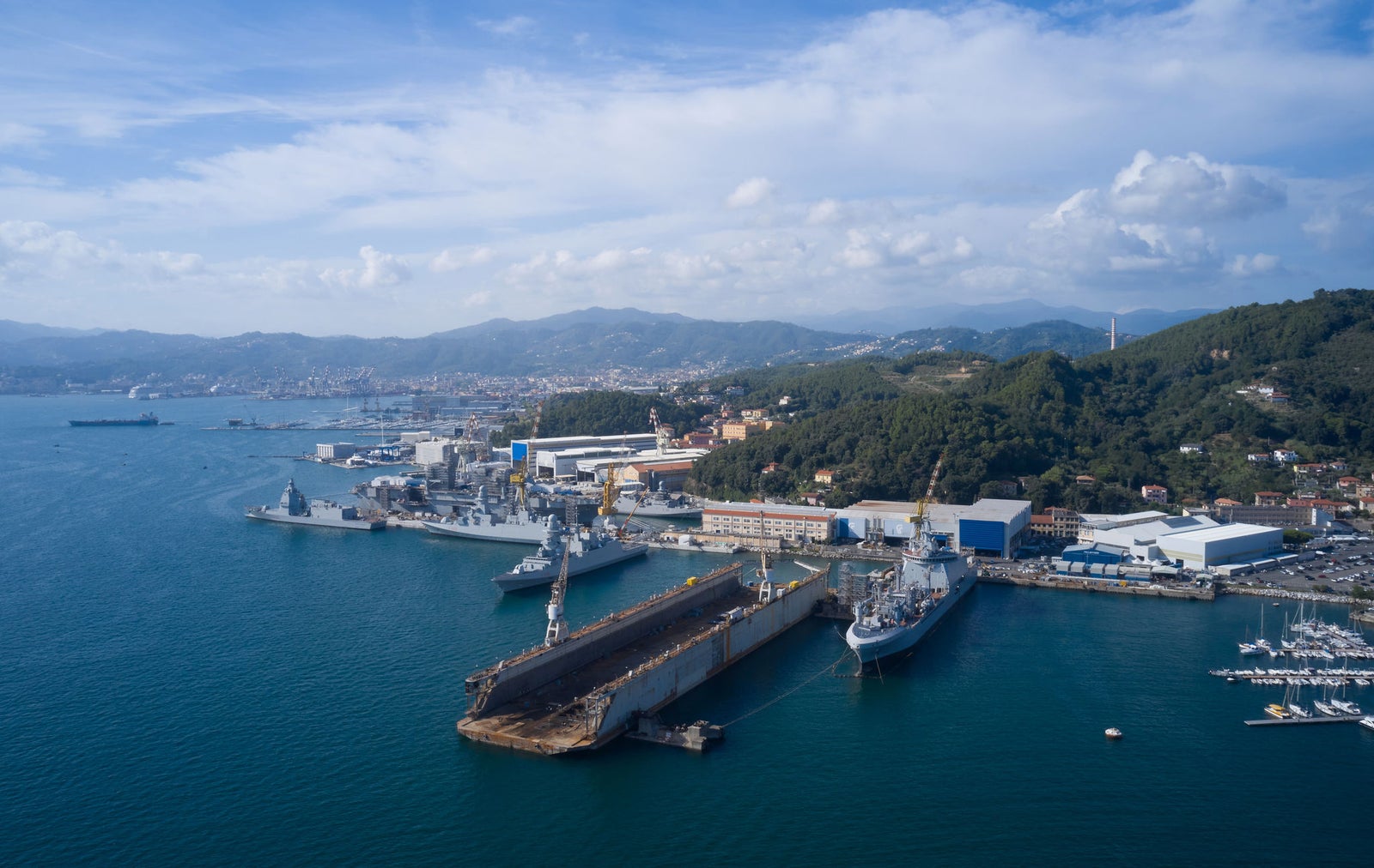 Fincantieri | Group - Sustainability - People and Community - Social Projects and Initiatives: Wide aerial view of a shipyard with many docked ships and a dry dock, coastal area between hills and industrial buildings, blue waters contrasting with green landscape, partly cloudy sky.