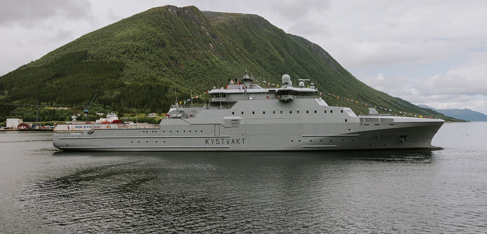 Fincantieri | Group - Business - Products - Defence Vessels - Patrol Vessels - KV Hopen: Side view of operational ship underway near coast, gray hull with "Kystvakt" text, visible antennas and equipment, green mountainous background and overcast sky.