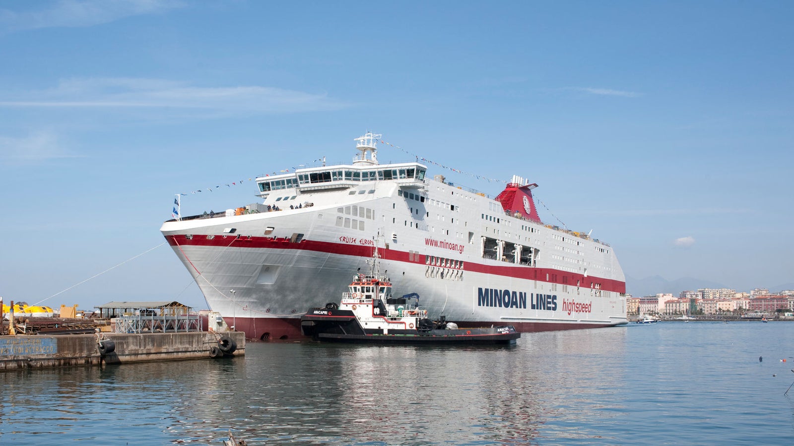 Fincantieri | Group - Business - Products - Ferries - Cruise Europa: Large Minoan Lines ferry docked at bustling port with smaller boats in foreground; calm waters and industrial structures present, clear blue sky overhead; visible text includes 'Minoan Lines' and 'Grimaldi Group', highlighting ferry branding.