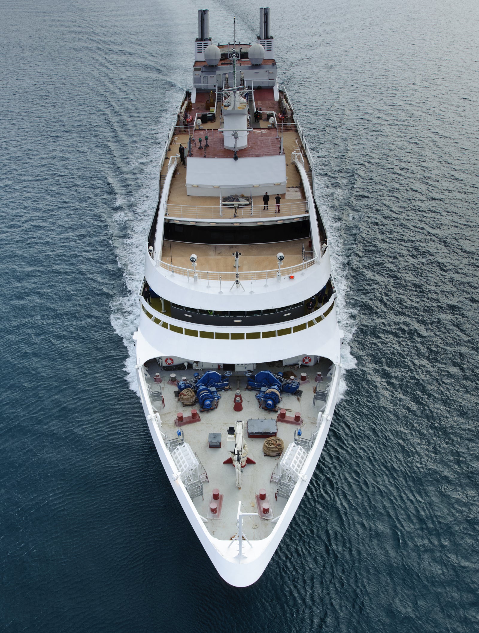 Fincantieri | Group - Business - Products - Cruise Ships - Le Boreal: Aerial view of Le Boreal cruise ship in operation navigating open sea, multiple passenger decks with relaxation areas and visible people, distant mountains and partly cloudy sky in background.