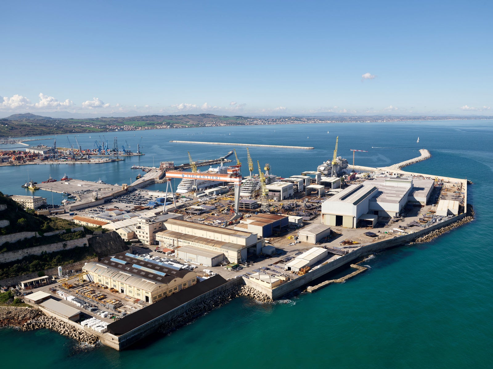 Fincantieri | Business Services - After-Sales Services - Lifecycle Management: An expansive shipyard and port facility captured from above, surrounded by calm blue waters. The image showcases large industrial buildings, cranes, and docked vessels, with a distant coastline under a clear sky. The setting highlights maritime infrastructure and coastal development.