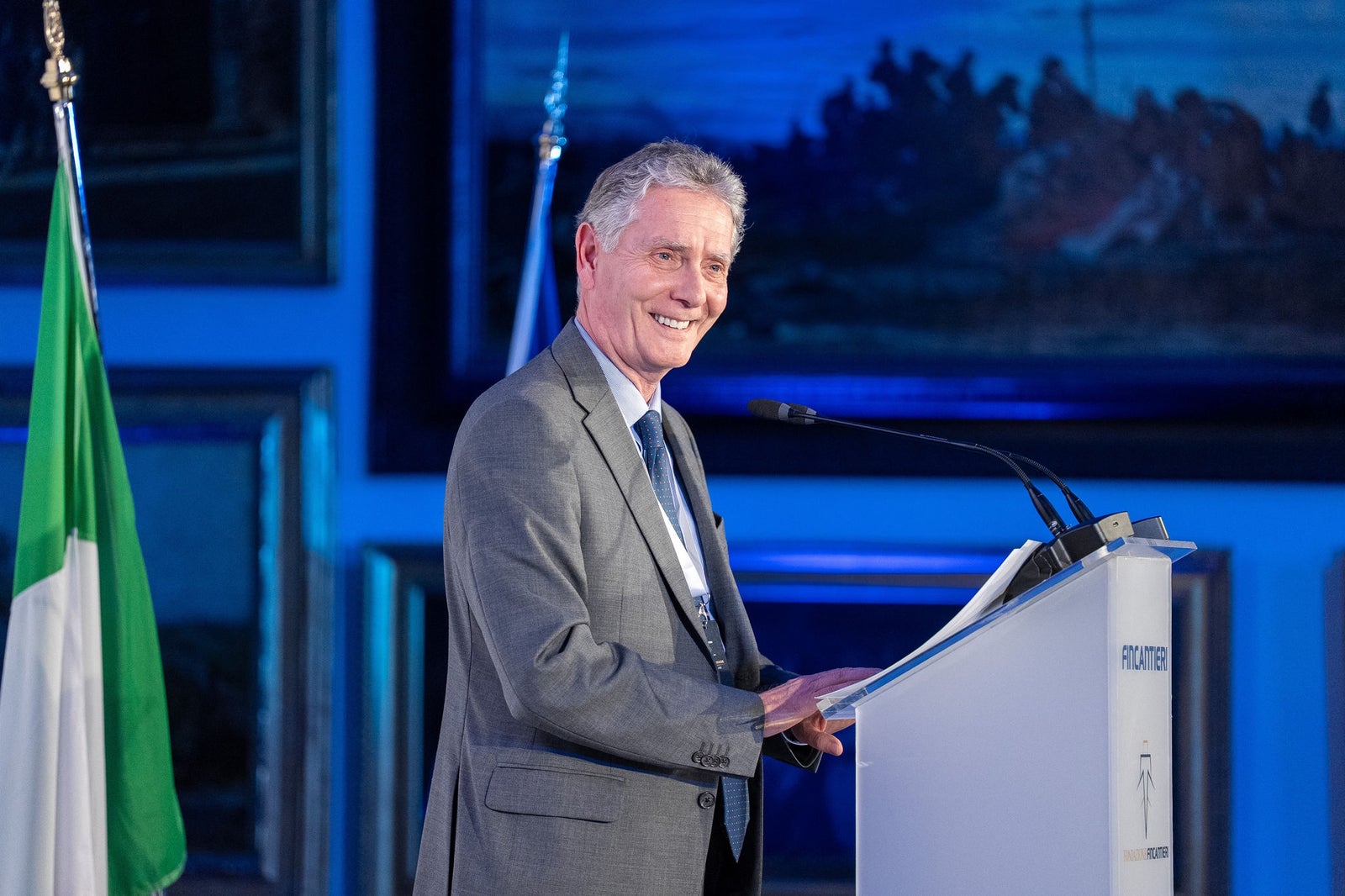 Newsroom - Comunicati stampa - 2025 - Gruppo Fincantieri: Un anno di Fondazione Fincantieri: storia, innovazione e impatto sociale: Relatore in abito istituzionale alla conferenza della Fondazione Fincantieri, ripreso di profilo al podio, con bandiera italiana e sfondo blu con quadri storici.