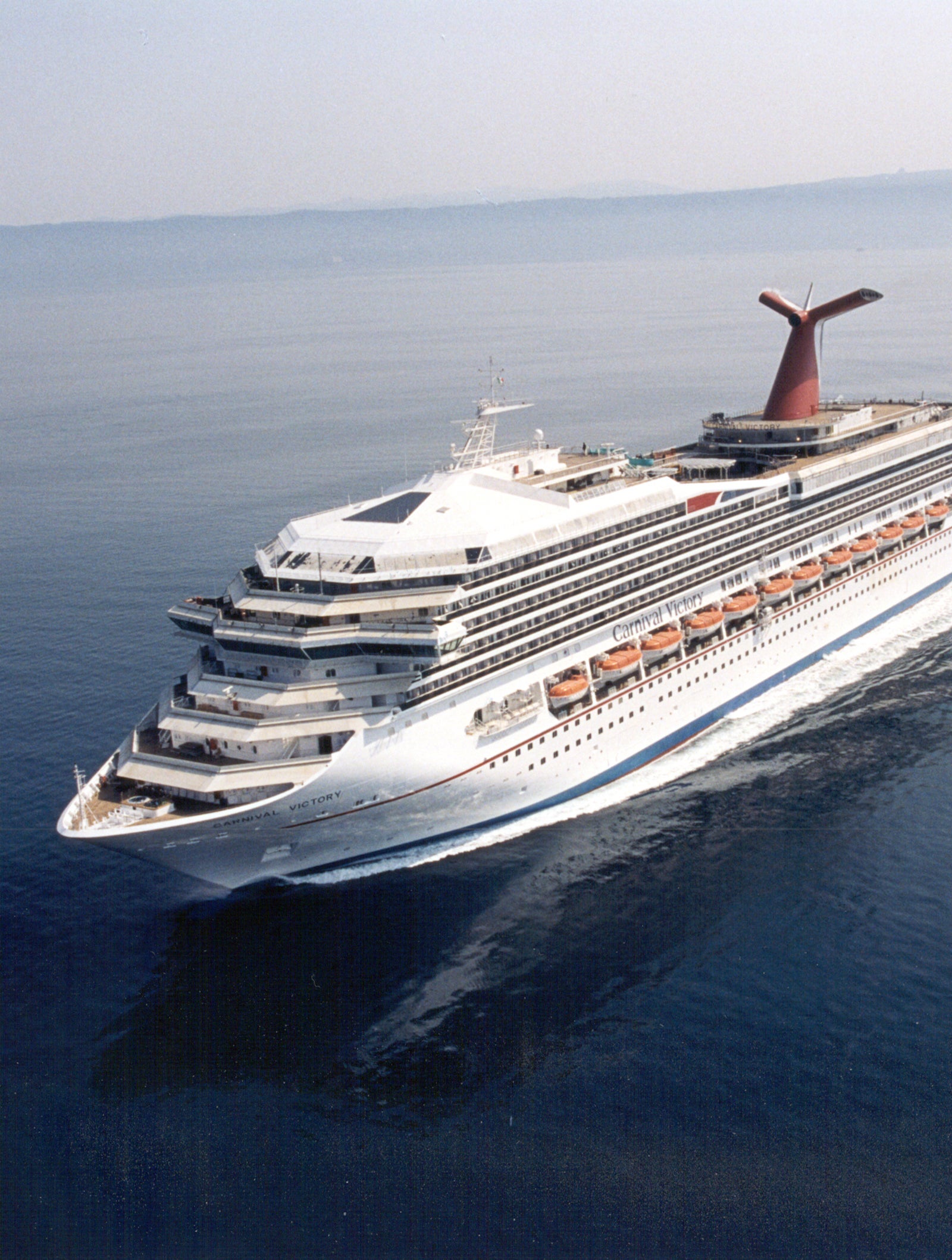 Fincantieri | Group - Business - Products - Cruise Ships - Carnival Victory: Large operational cruise ship gliding on calm open waters under clear sky, multiple decks with visible lifeboats, distinctive red funnel.