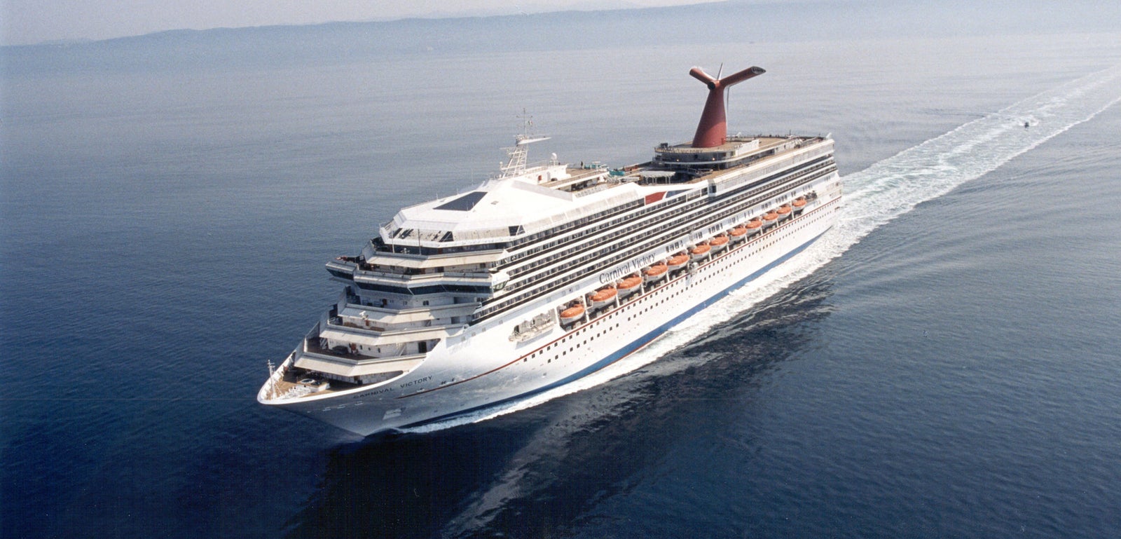 Fincantieri | Group - Business - Products - Cruise Ships - Carnival Victory: Large operational cruise ship gliding on calm open waters under clear sky, multiple decks with visible lifeboats, distinctive red funnel.