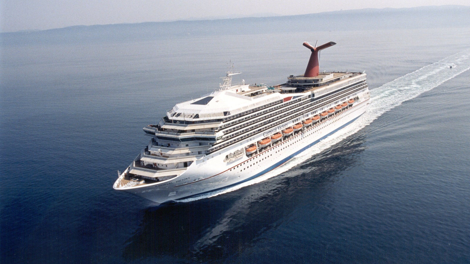 Fincantieri | Group - Business - Products - Cruise Ships - Carnival Victory: Large operational cruise ship gliding on calm open waters under clear sky, multiple decks with visible lifeboats, distinctive red funnel.