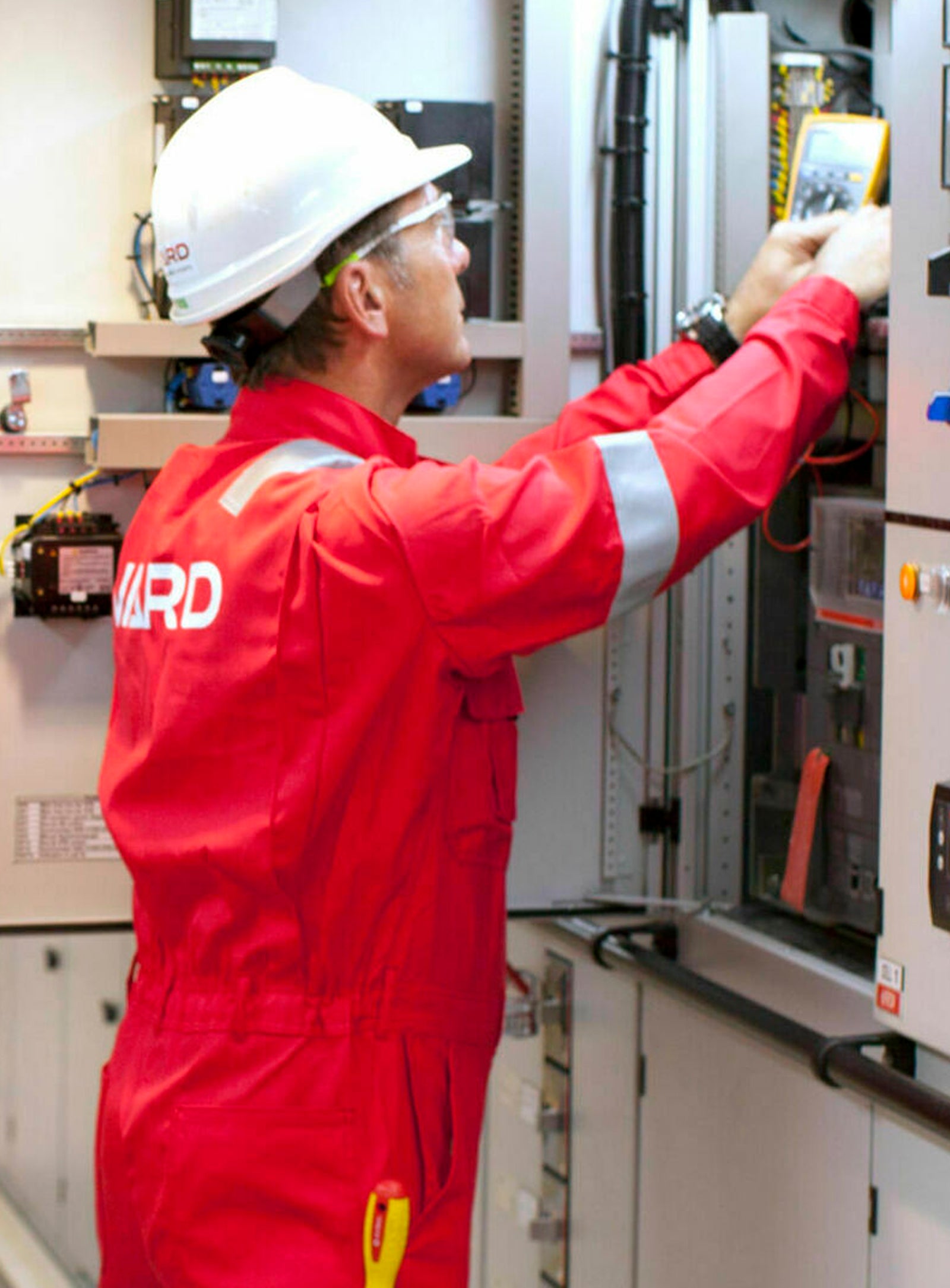 Fincantieri | Group - Subsidiaries and Affiliates - Vard Electro: Two technicians in red protective uniforms and white helmets working inside a control room, one operating a control panel and the other reviewing a document; industrial equipment, control panels, and structured workspace setting.