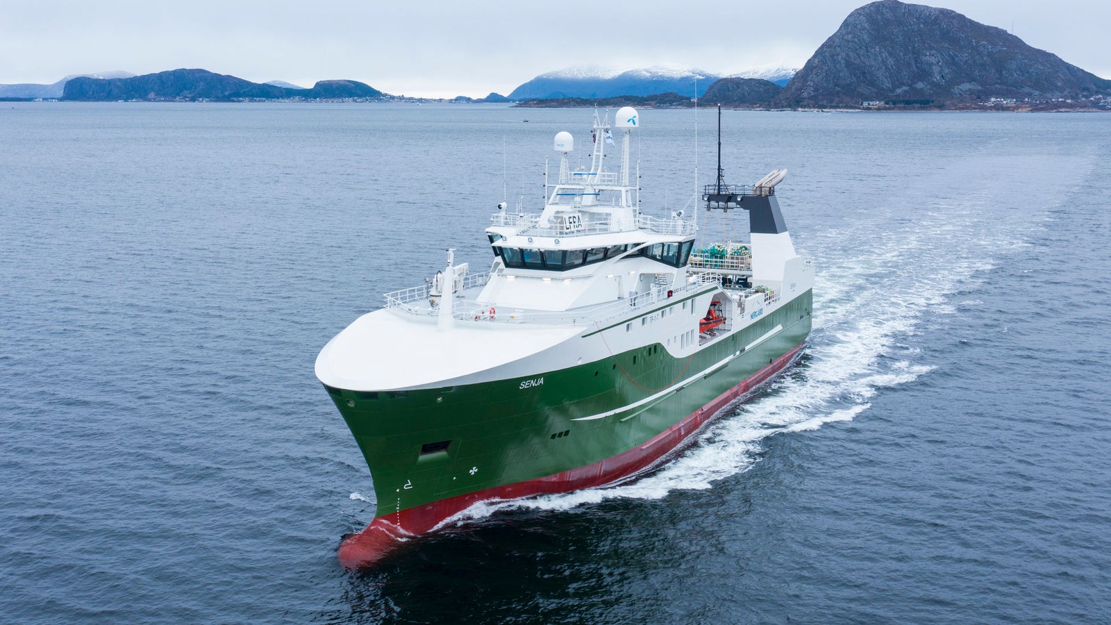 Prodotto - Peschereccio Senja: Vista laterale della nave operativa in navigazione, scafo verde e bianco, radar e sistemi di comunicazione avanzati, sfondo costiero montuoso, cielo coperto.